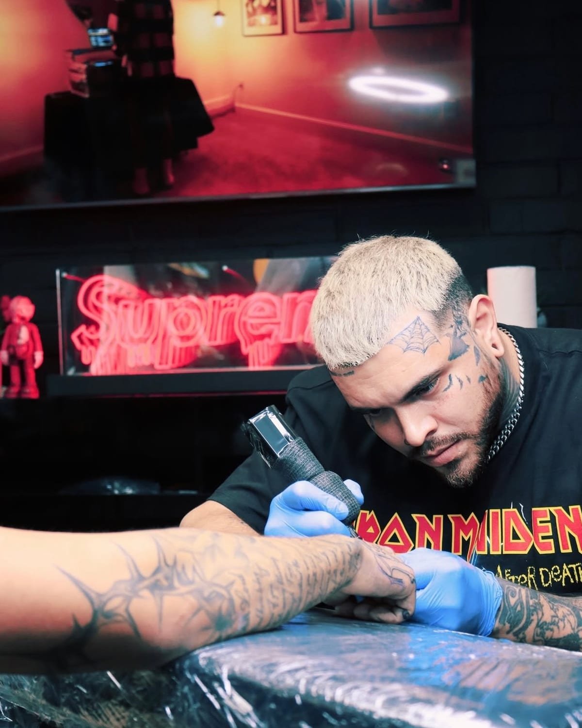 Santana leaning in over a forearm, shading with a tattoo machine under red neon.