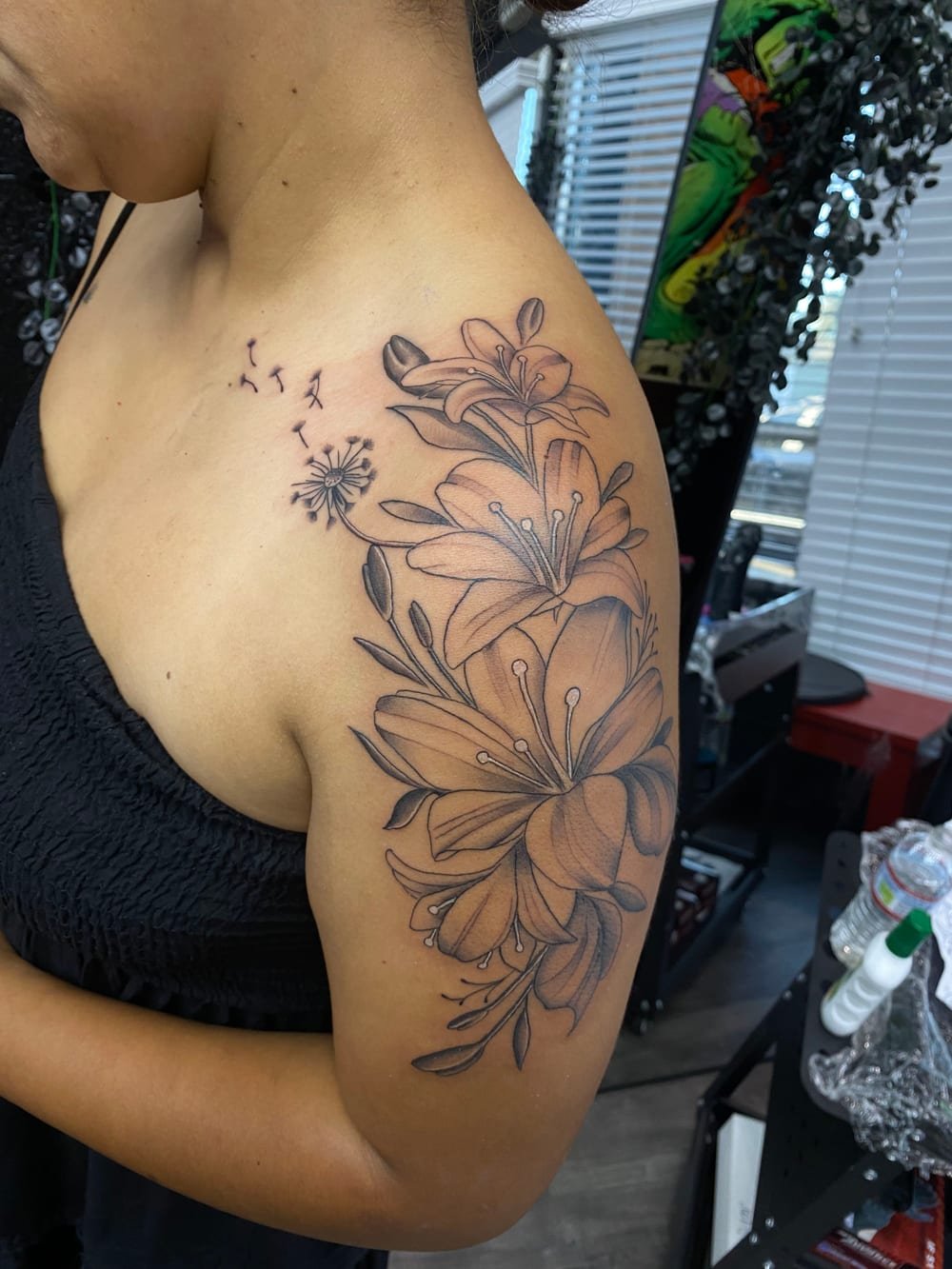 Fine-line black and grey lilies and dandelion seeds running across a shoulder and upper arm.