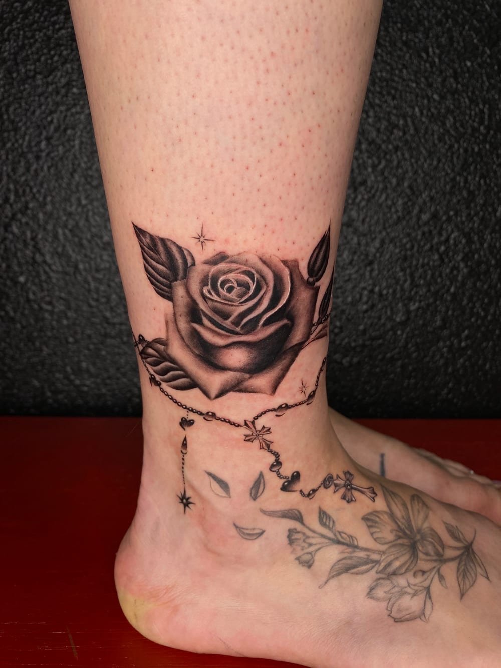 Fine-line black and grey rose with a rosary and trailing vines across an ankle and foot.
